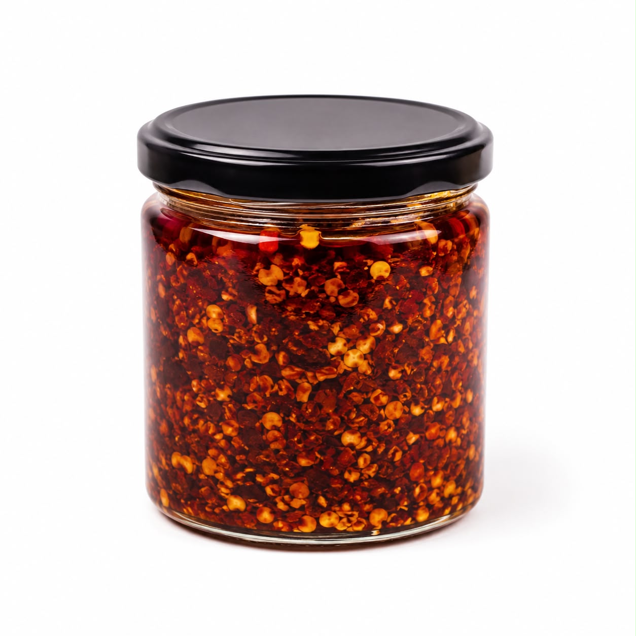 A clear glass jar filled with red chili pepper flakes in oil, sealed with a black lid, on a white background.