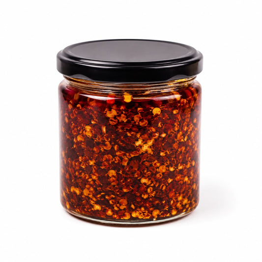 A clear glass jar filled with red chili pepper flakes in oil, sealed with a black lid, on a white background.