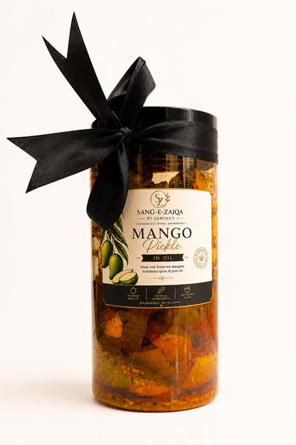 A glass jar of Sang-E-Zaiqa Mango Dinkle in Oil, filled with orange and yellow mango pieces, sealed with a black lid and decorated with a large black satin bow, set against a plain white background.